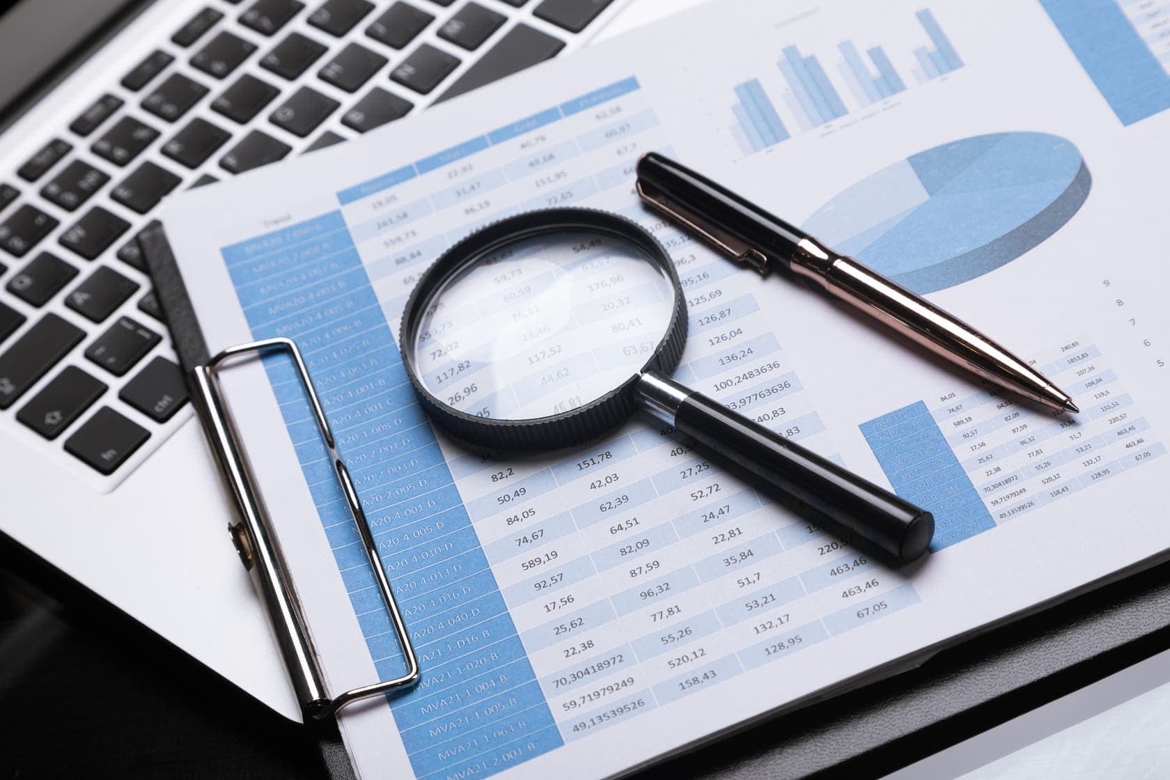 A magnifying glass and pen on a financial report beside a laptop